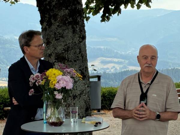 Quelle: Markus Weber Zwei Männer im Gespräch vor Publikum im Freien an einem Stehtisch mit Blumen, im Hintergrund der Stamm einer Linde und Aussicht auf den Schwarzwald.